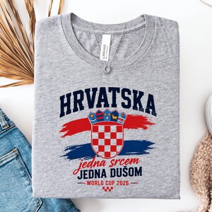 May include: A gray t-shirt with the word "HRVATSKA" in navy blue block letters above a Croatian coat of arms. Below the coat of arms is the text "jedna srcem JEDNA DU&Scaron;OM" in red script. The shirt also has "WORLD CUP 2026" printed on it.