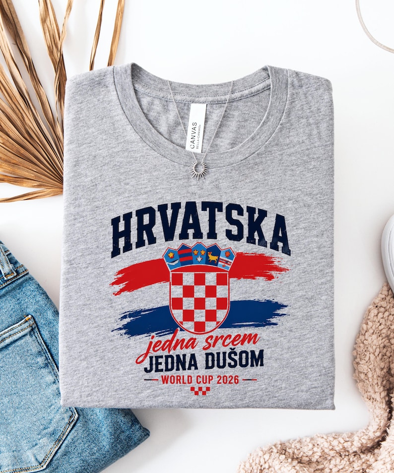 May include: A gray t-shirt with the word "HRVATSKA" in navy blue block letters above a Croatian coat of arms. Below the coat of arms is the text "jedna srcem JEDNA DU&Scaron;OM" in red script. The shirt also has "WORLD CUP 2026" printed on it.