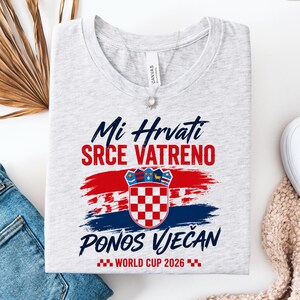 May include: A light gray t-shirt with the Croatian coat of arms and text in red and blue. The text reads "Mi Hrvati Srce Vatreno Ponos Vječan World Cup 2026". The design features red and blue brushstrokes.