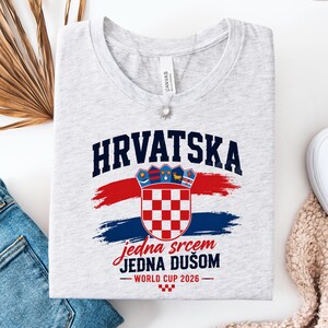 May include: A light gray t-shirt featuring the word "HRVATSKA" in navy blue, above a Croatian coat of arms and the text "jedna srcem JEDNA DU&Scaron;OM - WORLD CUP 2026" in red and blue. The shirt has a round neckline.