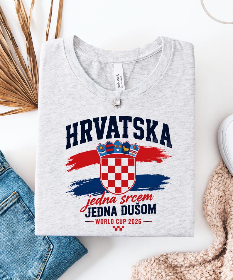 May include: A light gray t-shirt featuring the word "HRVATSKA" in navy blue, above a Croatian coat of arms and the text "jedna srcem JEDNA DU&Scaron;OM - WORLD CUP 2026" in red and blue. The shirt has a round neckline.