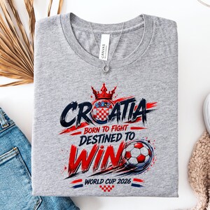 May include: A gray t-shirt with a graphic design featuring the word "CROATIA" in a bold, stylized font, along with the phrases "BORN TO FIGHT" and "DESTINED TO WIN." A soccer ball and the text "WORLD CUP 2026" are also included.