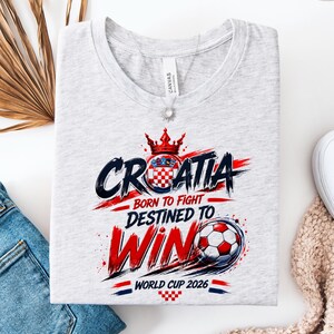 May include: A light gray t-shirt with a graphic design featuring the word "CROATIA" in black and red, a Croatian coat of arms, and the text "BORN TO FIGHT DESTINED TO WIN WORLD CUP 2026". A soccer ball is also featured.