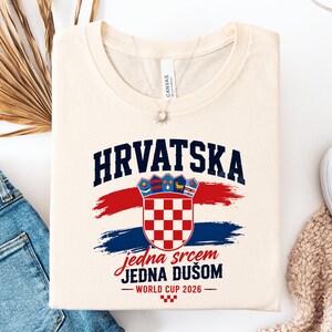 May include: Cream-colored t-shirt featuring the word "HRVATSKA" in navy blue above a Croatian coat of arms. Below the coat of arms are the words "jedna srcem JEDNA DU&Scaron;OM" in red script, with "WORLD CUP 2026" below.