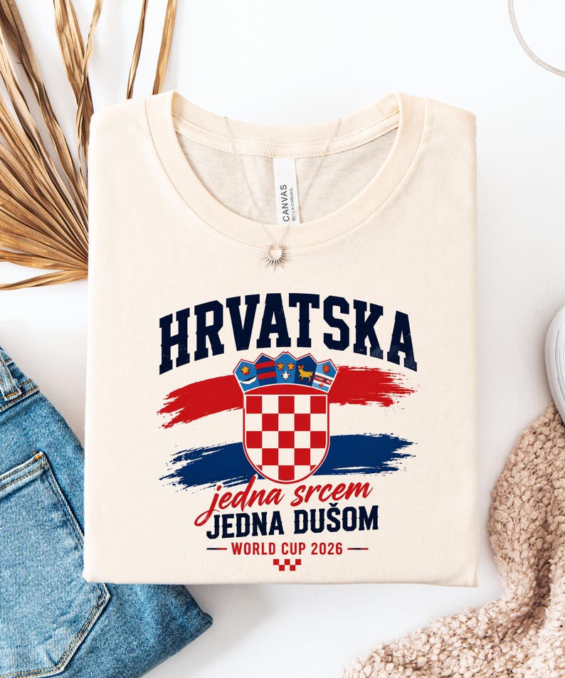May include: Cream-colored t-shirt featuring the word "HRVATSKA" in navy blue above a Croatian coat of arms. Below the coat of arms are the words "jedna srcem JEDNA DU&Scaron;OM" in red script, with "WORLD CUP 2026" below.