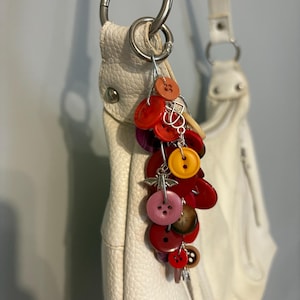 May include: A colorful button charm hangs from a white leather handbag. The charm features a cluster of various sized buttons in shades of red, orange, pink, yellow, and brown, connected by silver-toned metal. The handbag has silver hardware.
