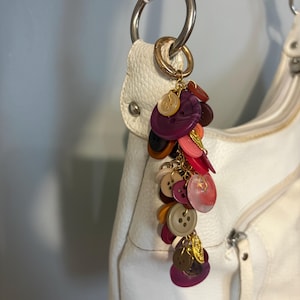 May include: A white leather handbag with a decorative charm. The charm features a cluster of colorful buttons in shades of purple, pink, brown, and beige, connected by a gold chain. The bag has silver hardware and a zipper.