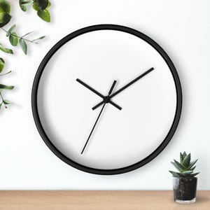 May include: A round wall clock with a white face and black hands and a black frame. The clock is set against a white wall, with a small potted plant and greenery in the background.