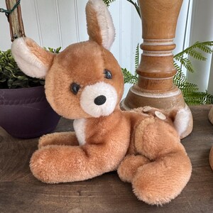 Puede incluir: Un juguete de peluche vintage con forma de ciervo en tonos marrones y blancos. El ciervo tiene ojos grandes y redondos, una nariz negra y orejas suaves y ca&iacute;das. Est&aacute; tumbado sobre una superficie de madera, con las patas estiradas. El juguete es un ejemplo cl&aacute;sico de peluche.