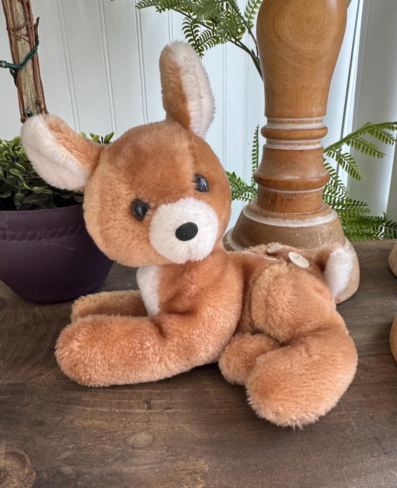 Puede incluir: Un juguete de peluche vintage con forma de ciervo en tonos marrones y blancos. El ciervo tiene ojos grandes y redondos, una nariz negra y orejas suaves y ca&iacute;das. Est&aacute; tumbado sobre una superficie de madera, con las patas estiradas. El juguete es un ejemplo cl&aacute;sico de peluche.