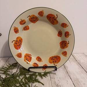 May include: A round ceramic bowl with a cream-colored interior and a dark green rim. The surface is decorated with multiple orange jack-o'-lantern pumpkin illustrations of varying sizes, each featuring a black hand-drawn face and stem.