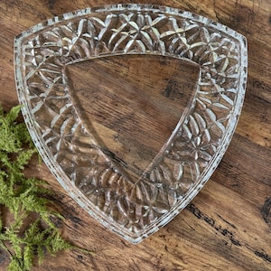 May include: Clear glass triangular serving dish with a detailed, textured pattern. The dish has a decorative edge and a smooth interior. The glass dish is displayed on a wooden surface, showcasing its design and clarity.