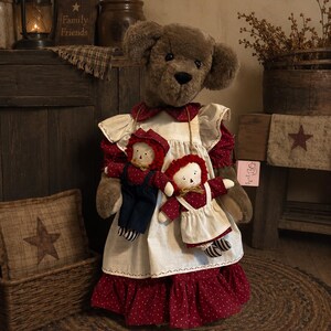May include: A plush brown teddy bear wearing a dark red dress with white polka dots and a white apron. The bear holds two smaller rag dolls, one in blue overalls and one in a matching red dress, both featuring stitched facial features and yarn hair.