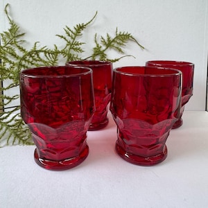May include: Four ruby red drinking glasses with a textured design. The glasses have a short, rounded base and a slightly flared rim. The glasses are arranged on a white surface with green foliage in the background.