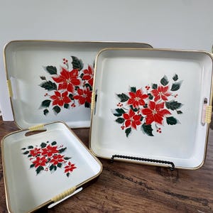 May include: Set of three white serving trays with gold trim and woven handles. Each tray features a poinsettia design with red flowers and green leaves. The trays are rectangular and square, perfect for serving food or displaying items.
