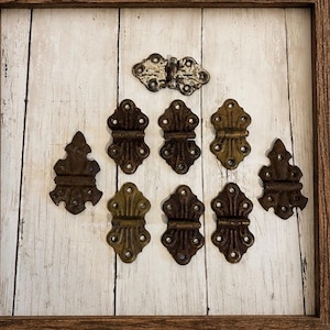 May include: A collection of nine metal hinges arranged in a grid. One hinge at the top is silver-toned, while the remaining eight hinges feature a dark, aged bronze finish. Each hinge has a decorative, symmetrical shape with multiple screw holes.