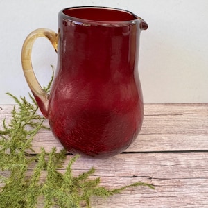May include: A ruby red glass pitcher with a textured surface and a tan handle. The pitcher has a spout for easy pouring. The handle is a contrasting color to the body of the pitcher. The pitcher is a decorative item.