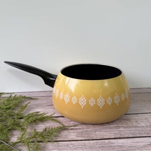 May include: A vintage yellow fondue pot with a black handle and interior. The pot features a white decorative pattern around the exterior. The pot is sitting on a wooden surface.