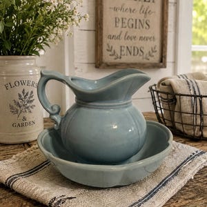 May include: A light blue ceramic pitcher and matching basin set. The pitcher features a curved handle and a rounded body, while the basin has a scalloped edge. Both pieces have a smooth, glossy finish and are resting on a textured fabric cloth.