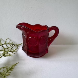 May include: A small, ruby-red glass creamer with a handle and a spout. The creamer has a decorative pattern of circles and a scalloped rim. The glass is a deep, transparent red, and the creamer is set against a white background.