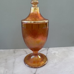 May include: An iridescent orange glass jar with a lid. The jar has a textured surface and a pedestal base. The lid is topped with a pointed finial. The jar is sitting on a marble surface.