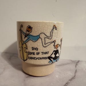 May include: A cream-colored ceramic mug with a crackle glaze finish. The exterior features a hand-drawn illustration of a musician playing a yellow upright bass and a person sitting, with the text "DIG SOME OF THAT CRAZYCOFFEE" printed in black.
