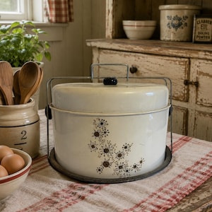 May include: A cream-colored metal cake carrier with a domed lid and a wire handle. The exterior features a dark brown starburst pattern. The carrier sits on a circular base and includes a small black knob on the top of the lid for easy lifting.