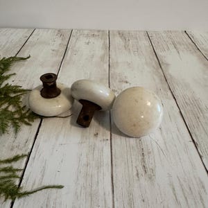 May include: Three round white porcelain door knobs with weathered brown metal shanks. Two of the knobs show the square metal spindle hole at the top of the shank, while the third knob is positioned to show its smooth, rounded surface.