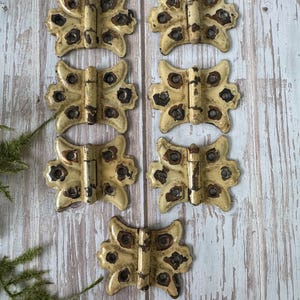 May include: Six antique butterfly-shaped hinges with a distressed, cream-colored finish and dark brown accents. The hinges are arranged on a light-colored, weathered wooden surface, showcasing their vintage aesthetic.