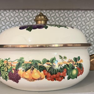 May include: Cream-colored enamelware casserole dish with a lid and gold-colored handles. The dish is decorated with a colorful fruit and vine design, including grapes, pears, and cherries. The lid is topped with a gold-colored finial.