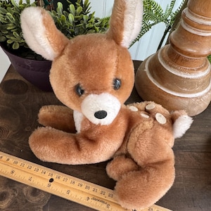 Puede incluir: Un juguete de peluche de ciervo marr&oacute;n con detalles blancos en las orejas, el pecho y la cola. El ciervo tiene ojos negros y est&aacute; tumbado. Una regla de madera con medidas en cent&iacute;metros est&aacute; en primer plano.