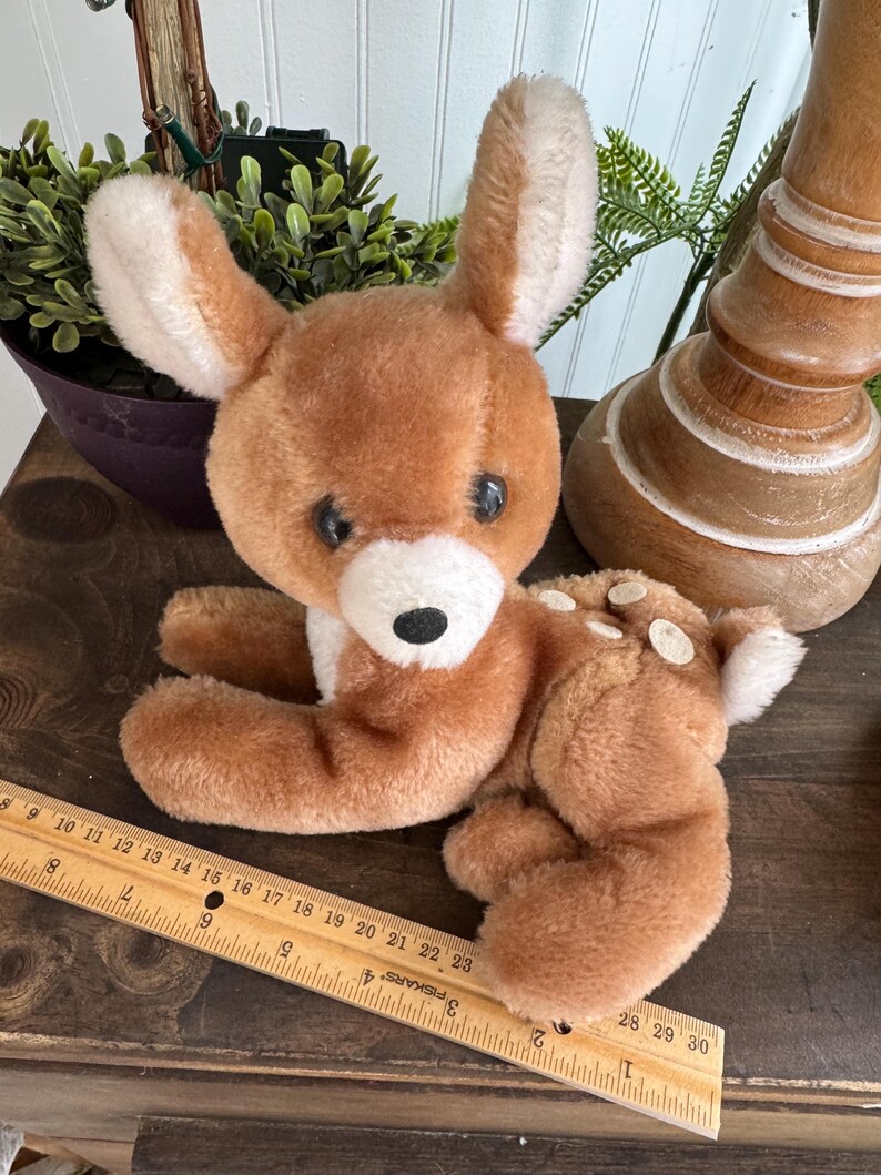 Puede incluir: Un juguete de peluche de ciervo marr&oacute;n con detalles blancos en las orejas, el pecho y la cola. El ciervo tiene ojos negros y est&aacute; tumbado. Una regla de madera con medidas en cent&iacute;metros est&aacute; en primer plano.