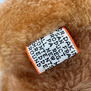 Puede incluir: Primer plano de una tela marr&oacute;n claro y texturizada. Se adjunta una peque&ntilde;a etiqueta rectangular con texto negro sobre fondo blanco. La etiqueta tiene un borde naranja y parece estar tejida.