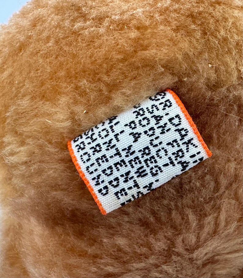 Puede incluir: Primer plano de una tela marr&oacute;n claro y texturizada. Se adjunta una peque&ntilde;a etiqueta rectangular con texto negro sobre fondo blanco. La etiqueta tiene un borde naranja y parece estar tejida.