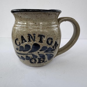 May include: A speckled stoneware mug with a dark rim and handle. The mug features the word "CANTON" in dark blue lettering, along with a decorative leaf design and the letters "OH". This handmade mug is perfect for coffee or tea.