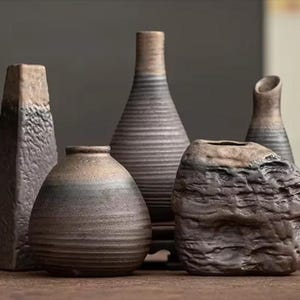 May include: A collection of five ceramic vases with dark, earthy tones. The set includes a tall rectangular vase, a rounded vase with horizontal ridges, a slender bottle-shaped vase, a small necked vase, and a textured, rock-like vessel.