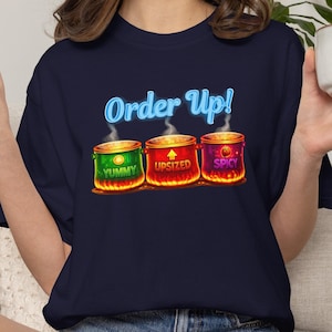 May include: Navy blue t-shirt with the text "Order Up!" in a light blue font. Below the text are three colorful pots with the words "Yummy," "Upsized," and "Spicy." The pots are green, red, and purple, respectively, with flames at the bottom.