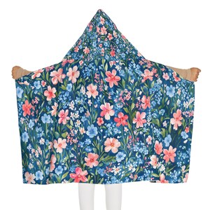 May include: A hooded blanket featuring a dark blue background with a vibrant floral pattern of pink, light blue, and white flowers with green leaves. The hooded garment displays a repeating botanical print across the entire fabric surface.