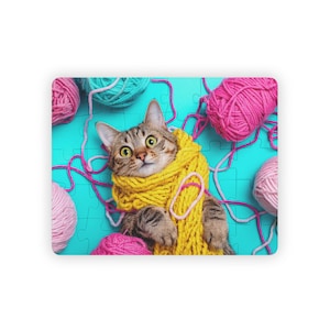 Könnte beinhalten: Eine getigerte Katze mit gelbem Schal, umgeben von rosa und türkisfarbenen Wollknäueln und Garnsträngen, auf einem türkisfarbenen Puzzle-Hintergrund. Die Katze hat große, überraschte Augen. Das Garn ist um die Katze herum angeordnet und erzeugt eine verspielte und farbenfrohe Szene.