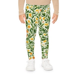 May include: Leggings featuring a white base with a repeating botanical pattern of dark green leaves and vibrant yellow flowers. The design covers the entire length of the leggings, which are worn with a beige long-sleeved top and white sneakers.
