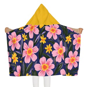 Pode incluir: Uma manta com capuz apresentando um capuz amarelo mostarda sólido e uma base azul-marinho escuro. O tecido é decorado com um padrão repetitivo de grandes flores rosa claro com centros amarelos, flores amarelas menores e folhas verde-escuras.