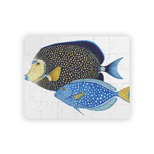 Könnte beinhalten: Ein Puzzlespiel mit zwei farbenfrohen Fischillustrationen. Der größere Fisch ist dunkelblau mit gelben Punkten, einem gelben Gesicht und einer blauen Flosse. Der kleinere Fisch ist blau mit weißen Punkten und einer gelben Schwanzflosse.