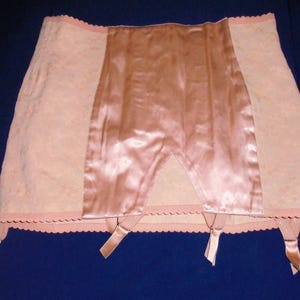 Pode incluir: Um cinto de ligas de cintura alta na cor pêssego, com um painel central de cetim liso e brilhante, ladeado por dois painéis laterais de tecido texturizado com padrão. A borda inferior tem um acabamento recortado e quatro tiras de liga.