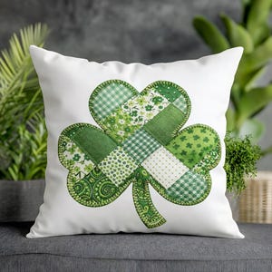 May include: White square pillow featuring a large, green patchwork shamrock design. The shamrock is made of various green fabrics with different patterns, including floral, gingham, and solid textures. The edges are outlined with green stitching.