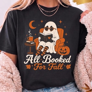 Puede incluir: Una camiseta negra con el gráfico de un fantasma con gafas de sol en forma de corazón, sentado en un sillón naranja leyendo un libro. El diseño incluye una calabaza, una taza de café, una luna creciente y el texto "All Booked For Fall" en blanco y naranja.