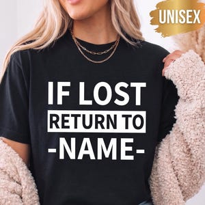 Könnte beinhalten: Ein schwarzes kurzärmeliges T-Shirt mit dem weißen Text IF LOST RETURN TO -NAME- auf der Vorderseite. Ein goldfarbenes grafisches Abzeichen mit dem Wort UNISEX befindet sich in der oberen rechten Ecke des Bildes.