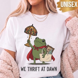 May include: A white short-sleeve t-shirt featuring a graphic of a green frog wearing a red cape, holding a stained-glass lamp, and carrying a tote bag with a picture frame and bottle. The text "WE THRIFT AT DAWN" is printed below the frog graphic.