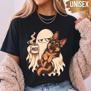 Może przedstawiać: Czarny t-shirt z grafiką przedstawiającą białego ducha trzymającego kubek kawy z napisem MEH, obejmującego brązowo-czarnego szczeniaka owczarka niemieckiego. Koszulka zawiera złotą etykietę graficzną z napisem UNISEX.