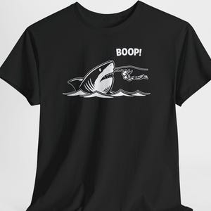 Può includere: Una t-shirt nera a maniche corte con una grafica bianca di uno squalo che emerge dalle onde. Una mano si allunga per toccare il naso dello squalo, accompagnata dalla scritta bianca BOOP! stampata sopra lo squalo. Il disegno è centrato.