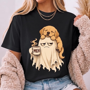 Könnte beinhalten: Ein schwarzes T-Shirt mit einem Grafikdruck eines beigen Geistes mit mürrischem Gesichtsausdruck, der eine mit MEH beschriftete Tasse hält, wobei ein Golden Retriever Welpe auf dem Kopf des Geistes ruht. Das Motiv ist vorne auf dem T-Shirt zentriert.